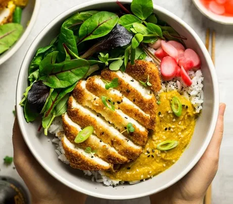 A bowl with breaded tofu, greens, radishes, curry sauce, and rice, garnished with sesame seeds and green onions.