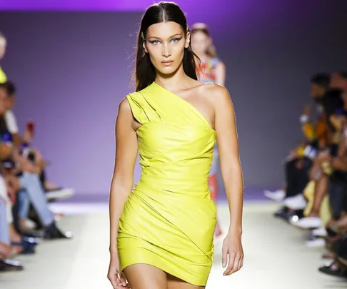 Model in a bright yellow one-shoulder leather outfit walking down a runway with a focused expression.