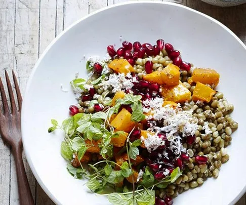 A colorful salad with lentils, pomegranate seeds, squash, greens, and shredded coconut on a white plate with wooden utensils nearby.