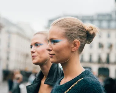 Two women wearing blue eyeshadow walk outdoors, with hair styled in buns, against a blurred city background.