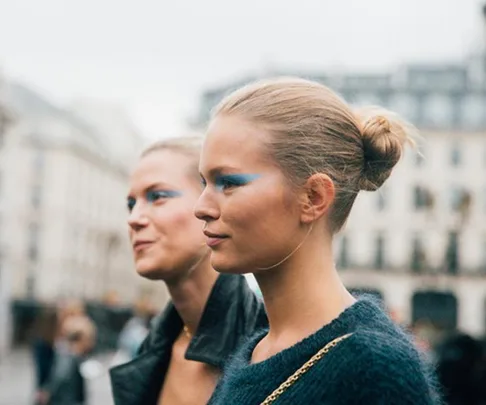 Two women wearing blue eyeshadow walk outdoors, with hair styled in buns, against a blurred city background.