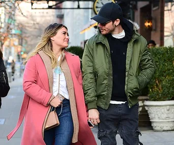 A couple strolling down a city sidewalk, the woman in a pink coat and the man in a green jacket and black cap, holding hands.