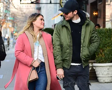 A couple strolling down a city sidewalk, the woman in a pink coat and the man in a green jacket and black cap, holding hands.