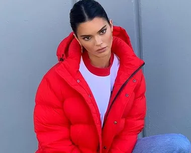 Kendall Jenner poses in a red puffer jacket, white t-shirt, and blue jeans, with a neutral expression against a grey wall.