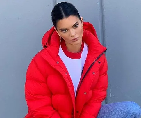 Kendall Jenner poses in a red puffer jacket, white t-shirt, and blue jeans, with a neutral expression against a grey wall.