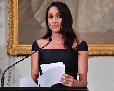 Meghan Markle delivering a speech in New Zealand, holding papers and standing at a podium with a microphone.