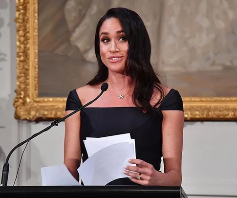 Meghan Markle delivering a speech in New Zealand, holding papers and standing at a podium with a microphone.