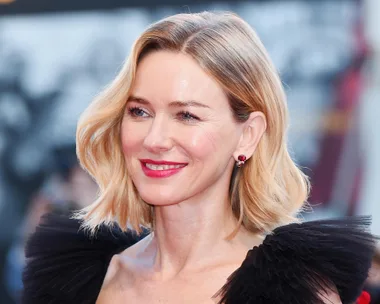 Naomi Watts at a red carpet event, wearing a black dress with sheer ruffles and red earrings.