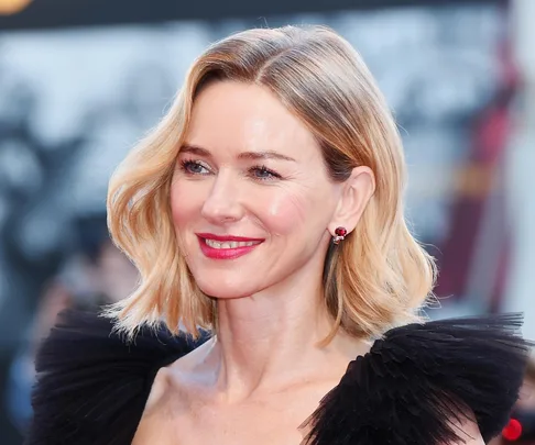 Naomi Watts at a red carpet event, wearing a black dress with sheer ruffles and red earrings.