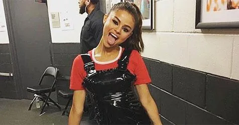 Young woman in a red top and black overalls playfully sticking out her tongue backstage.