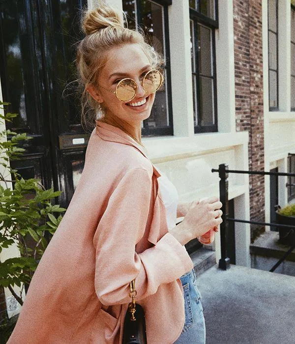 A woman in round sunglasses and a pink jacket smiles, standing on a sidewalk near a brick building.