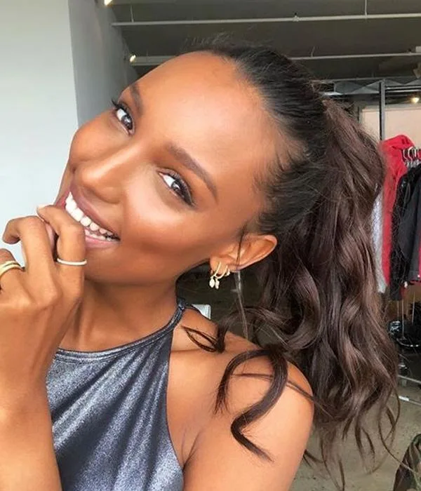 A woman with long curly hair smiles while gently touching her chin, wearing a shiny sleeveless top.
