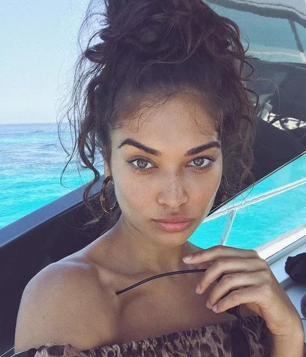 Woman with curly hair on a boat, holding sunglasses, with the ocean in the background.