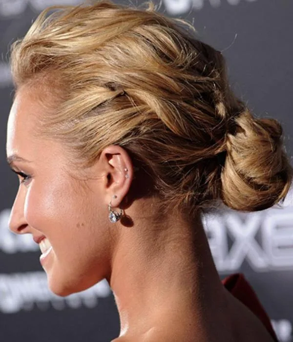 Profile of woman with blonde hair in a bun, wearing earrings, against a blurred dark background.