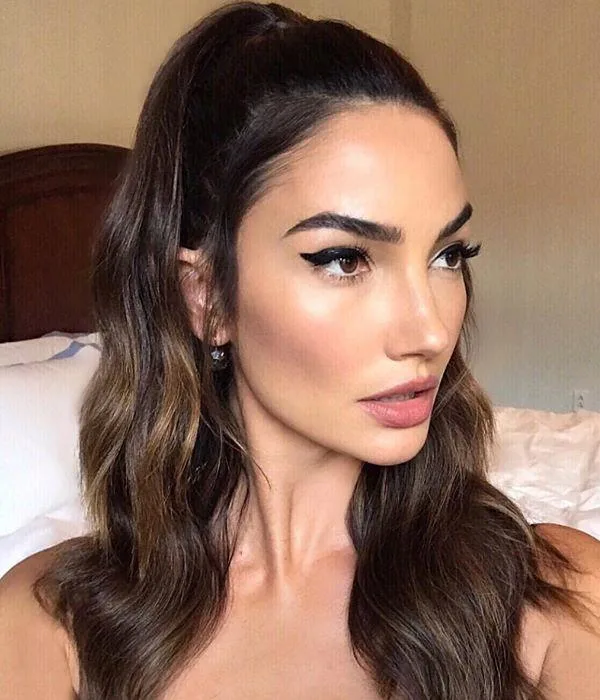 A woman with long brown hair and winged eyeliner, looking to the side, in a softly lit room.
