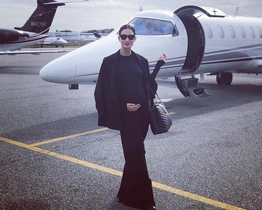 A woman in black clothing stands in front of a private jet with an open door, holding a bag.