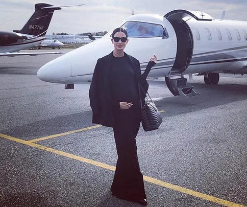 A woman in black clothing stands in front of a private jet with an open door, holding a bag.