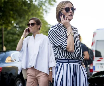 Two stylish women talking on their cell phones outdoors; one in striped attire and the other in white top with pink pants.