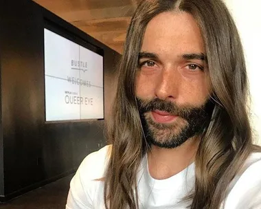 Jonathan Van Ness with long hair and a beard, wearing a white shirt, standing indoors with a "Queer Eye" sign in the background.