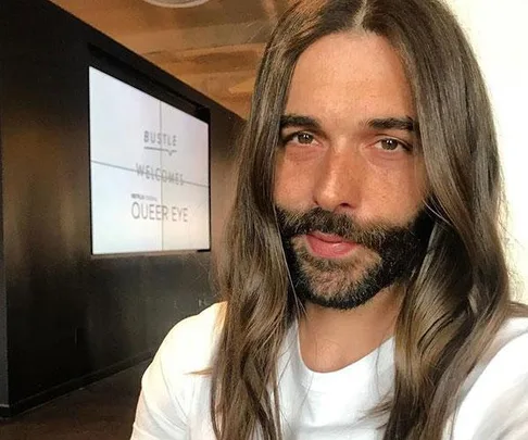 Jonathan Van Ness with long hair and a beard, wearing a white shirt, standing indoors with a "Queer Eye" sign in the background.