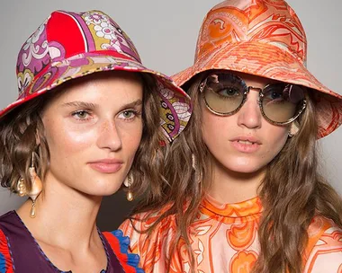 Two models in vibrant, patterned bucket hats. One wears sunglasses, and both don stylish, colorful outfits.