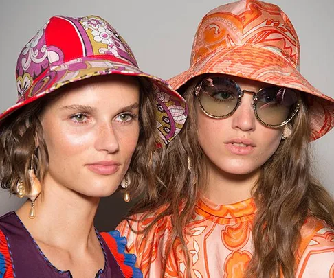 Two models in vibrant, patterned bucket hats. One wears sunglasses, and both don stylish, colorful outfits.