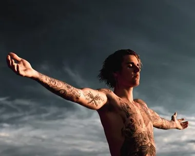 Shirtless man with arms outstretched, displaying numerous tattoos against a cloudy sky backdrop.