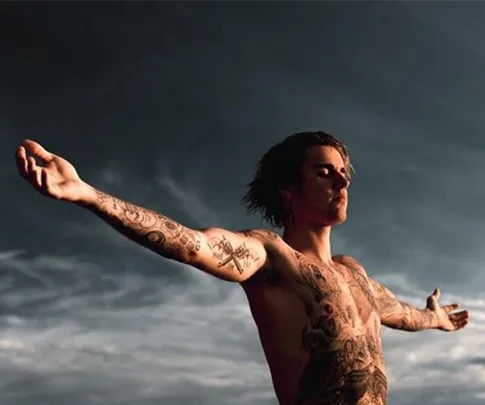 Shirtless man with arms outstretched, displaying numerous tattoos against a cloudy sky backdrop.