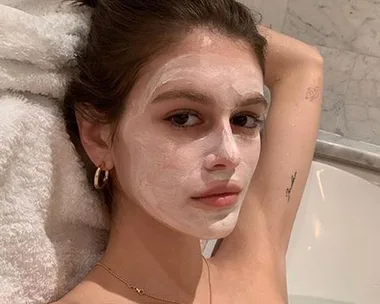 Woman in a bathtub with a white face mask, wearing gold hoop earrings and a necklace.