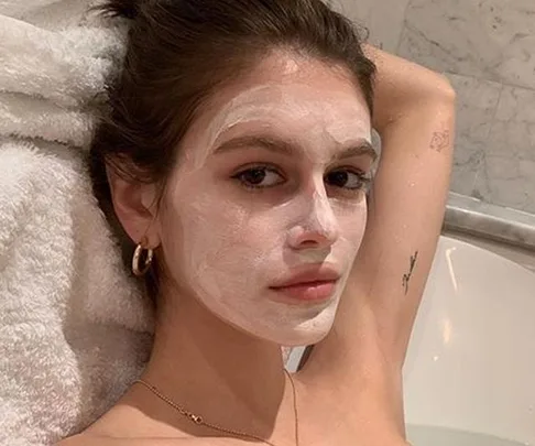 Woman in a bathtub with a white face mask, wearing gold hoop earrings and a necklace.