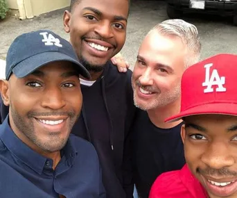 Four men smiling for a selfie, two wearing LA caps, in front of a beige building and black car.