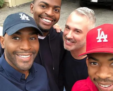 Four men smiling for a selfie, two wearing LA caps, in front of a beige building and black car.