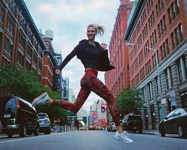 A person wearing athletic clothing joyfully jumps in the middle of a city street lined with red-brick buildings.