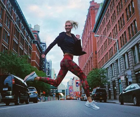 A person wearing athletic clothing joyfully jumps in the middle of a city street lined with red-brick buildings.