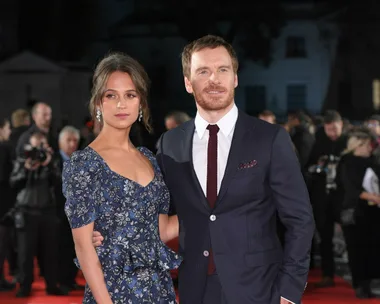 Two people, man in suit and woman in floral dress, pose on the red carpet with photographers in the background.