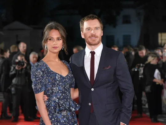 Two people, man in suit and woman in floral dress, pose on the red carpet with photographers in the background.