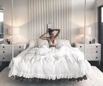 Woman sitting on a white bed in a bright, stylish bedroom with two side lamps and modern decor.