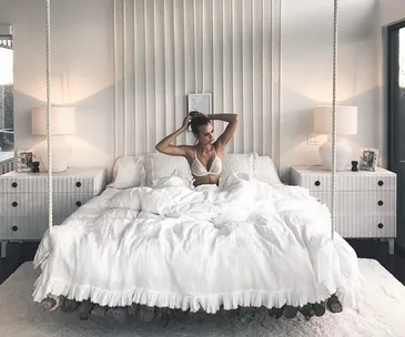 Woman sitting on a white bed in a bright, stylish bedroom with two side lamps and modern decor.