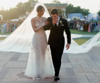 Bride in a lace gown and groom in a tux walk arm-in-arm outdoors, surrounded by guests.