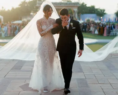 Bride in a lace gown and groom in a tux walk arm-in-arm outdoors, surrounded by guests.