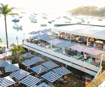 Rooftop bar with striped umbrellas overlooking a sunny harbor filled with boats.