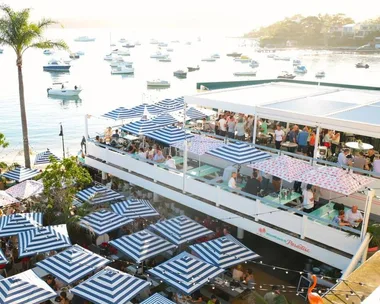 Rooftop bar with striped umbrellas overlooking a sunny harbor filled with boats.