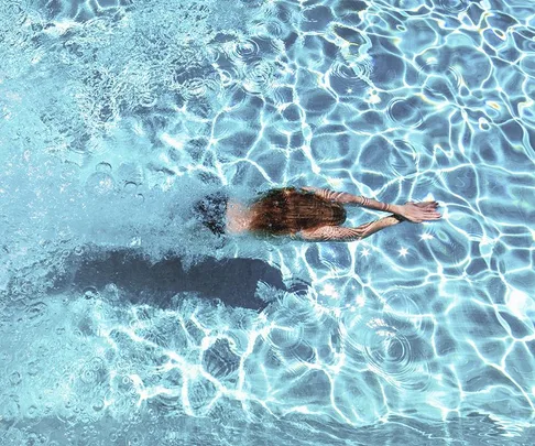 Person swimming underwater in a clear blue pool with sunlight creating ripple patterns on the surface.