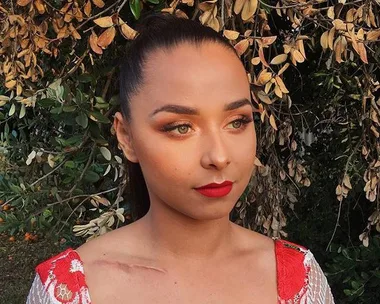 Person with makeup and red lipstick, wearing a white and red dress, posing outside with dry leaves in the background.