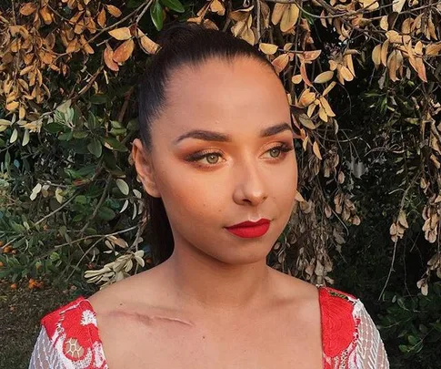 Person with makeup and red lipstick, wearing a white and red dress, posing outside with dry leaves in the background.