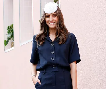 A woman in a navy outfit with a white hat smiles against a light pink wall at a spring racing event.