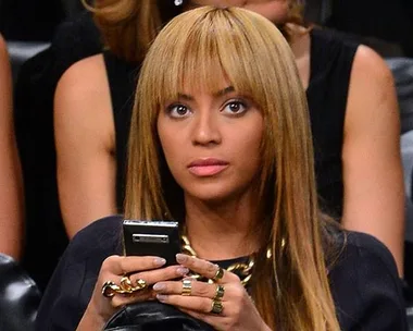 A woman with long, light brown hair and bangs, wearing statement rings, looks up while holding a phone.