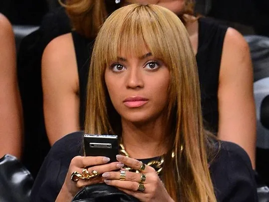 A woman with long, light brown hair and bangs, wearing statement rings, looks up while holding a phone.