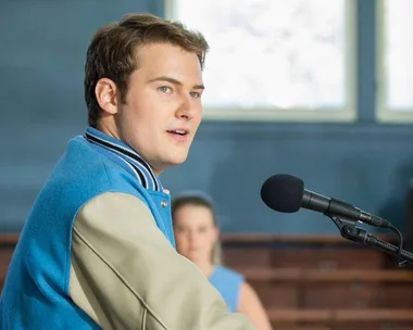 Young man in a varsity jacket speaks into a microphone in a gymnasium, from the series "13 Reasons Why".