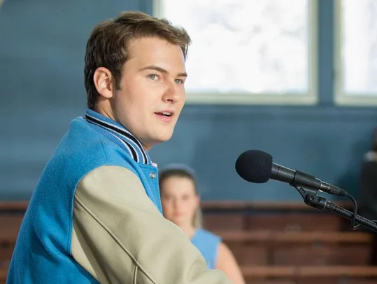 Young man in a varsity jacket speaks into a microphone in a gymnasium, from the series "13 Reasons Why".